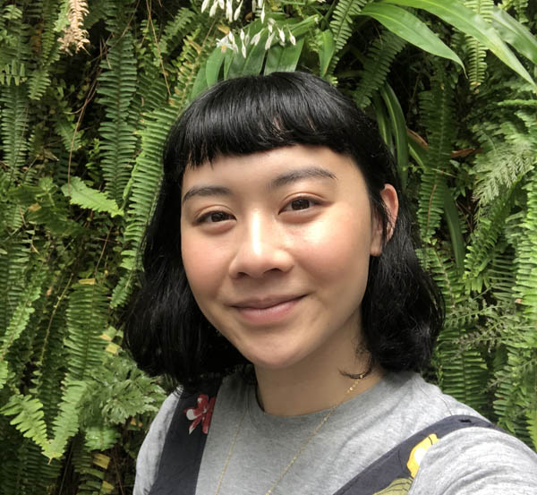 Artist bio image: Kim Anderson Kim stands in front of a wall of ferns smiling gently at the camera.